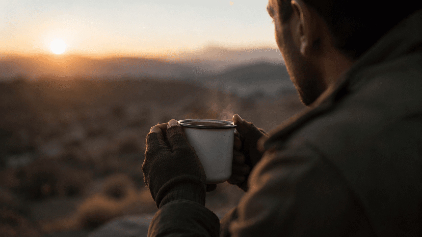 A soldier in dark fatigues holding a steaming coffee mug, looking toward the sunrise