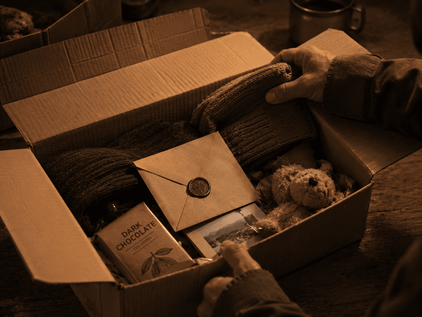 Hands in olive fatigues opening a cardboard care package containing folded clothing, chocolate, a knit cap, and a sealed envelope